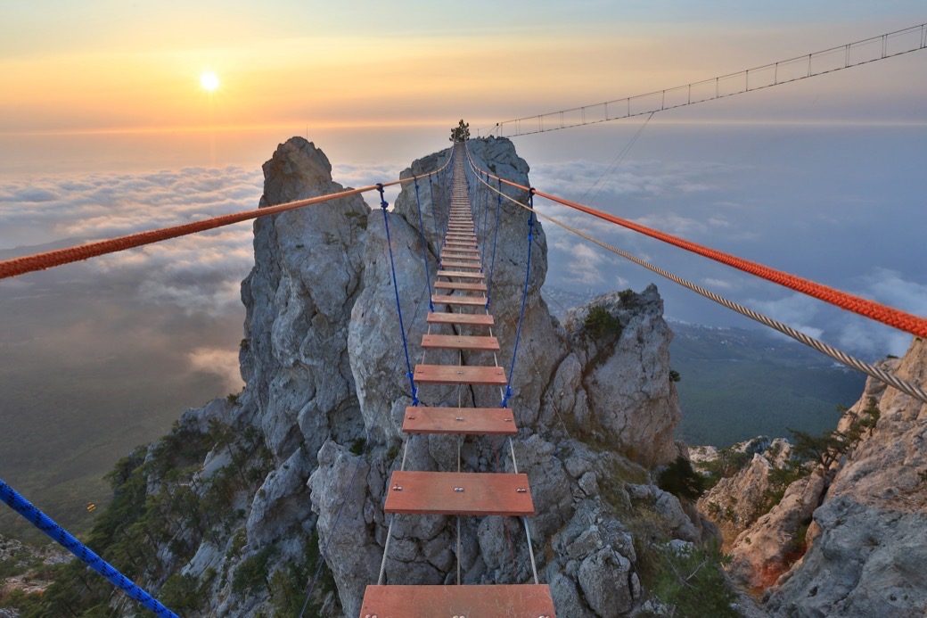 A rope bridge crossing between two peaks above the clouds at sunrise