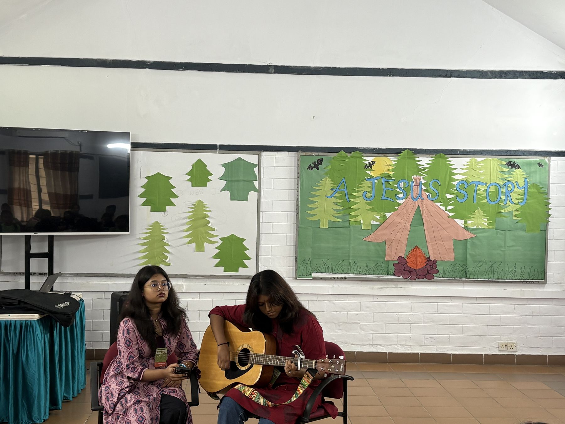Two young women leading songs with a guitar in front of a paper forest mural